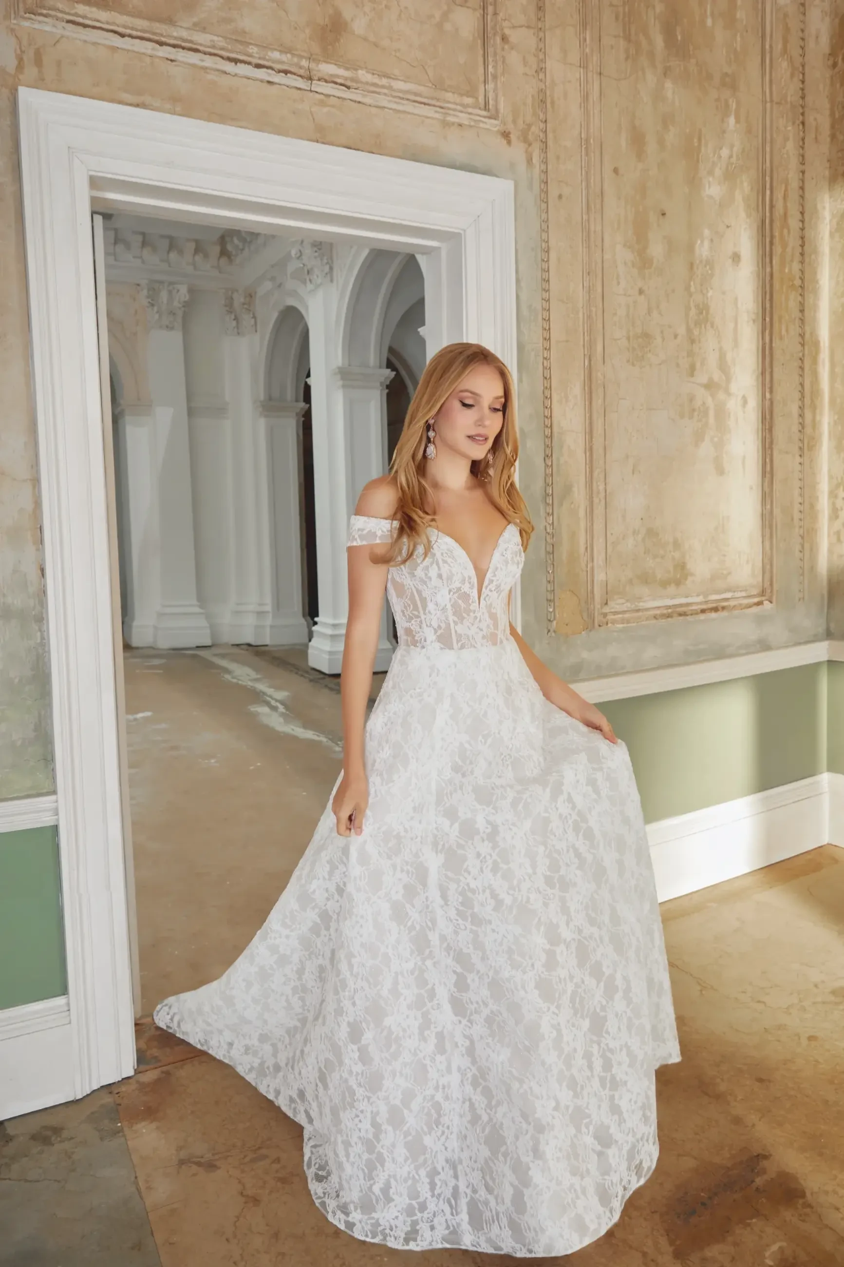 A woman in a white lace off-the-shoulder gown stands gracefully in an elegant, vintage room with textured walls and a grand archway.