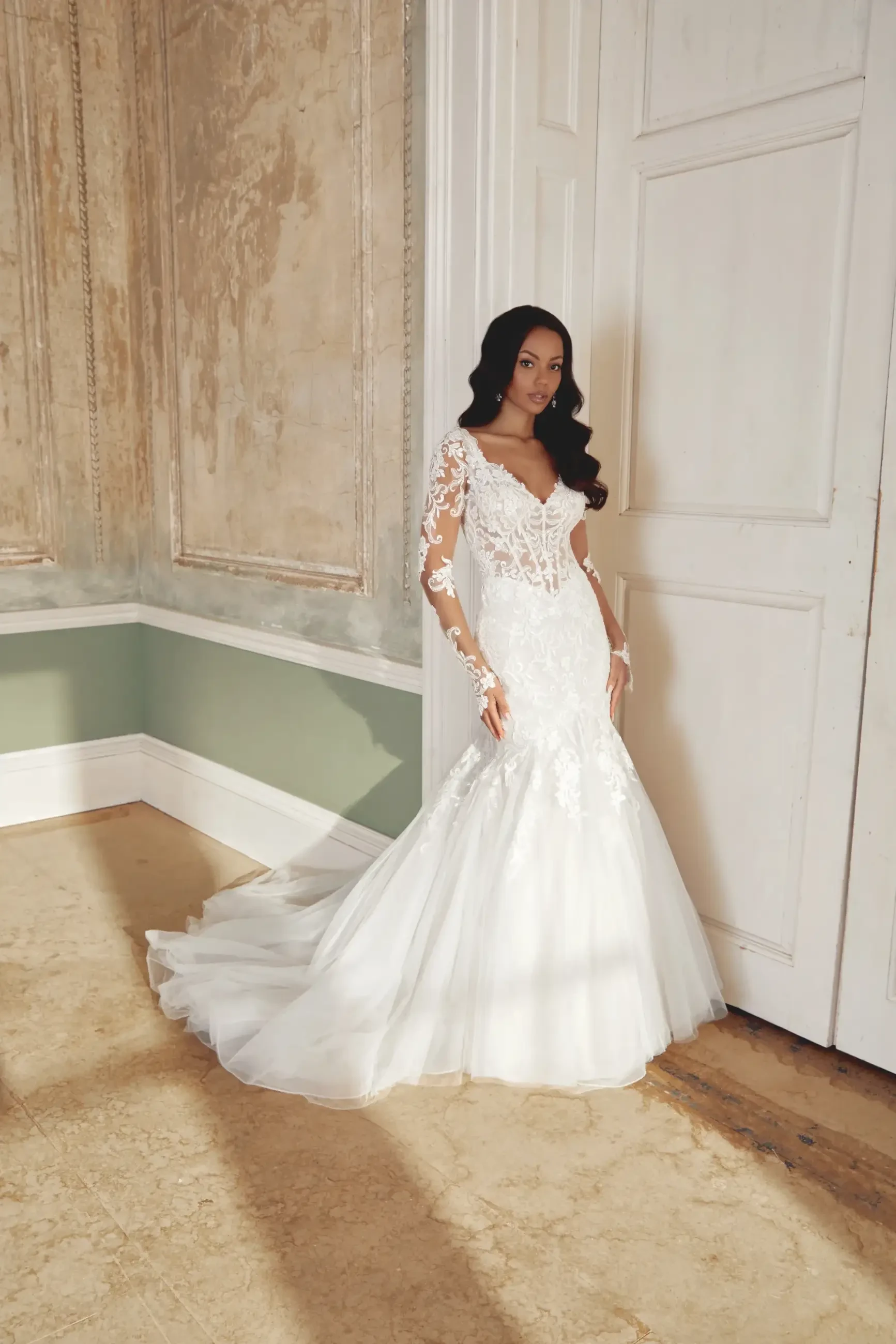 A bride in a detailed lace wedding gown with a flowing train stands elegantly by a white door in an ornate room, conveying elegance and grace.