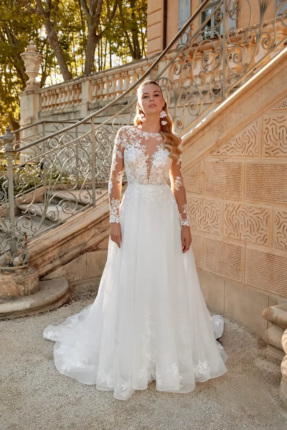 A woman in a white wedding dress with long lace sleeves, standing on a staircase in a picturesque outdoor setting.