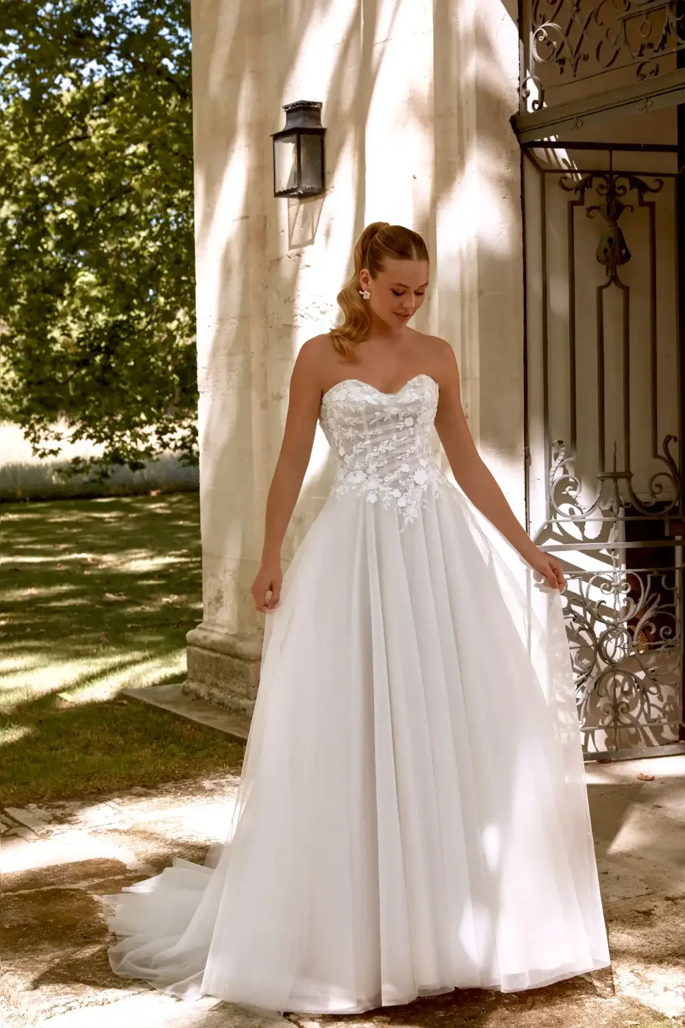 A smiling woman in a strapless white wedding dress with a flowing skirt, standing outdoors near a stone wall and vintage lanterns.
