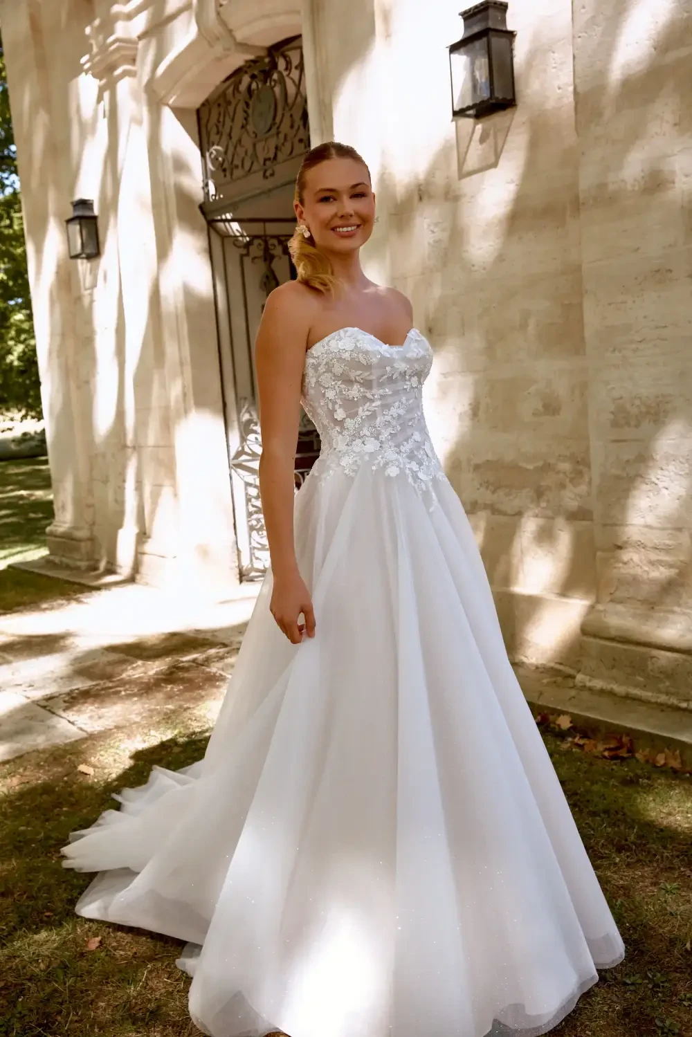 A smiling woman in a strapless white wedding dress with a flowing skirt, standing outdoors near a stone wall and vintage lanterns.