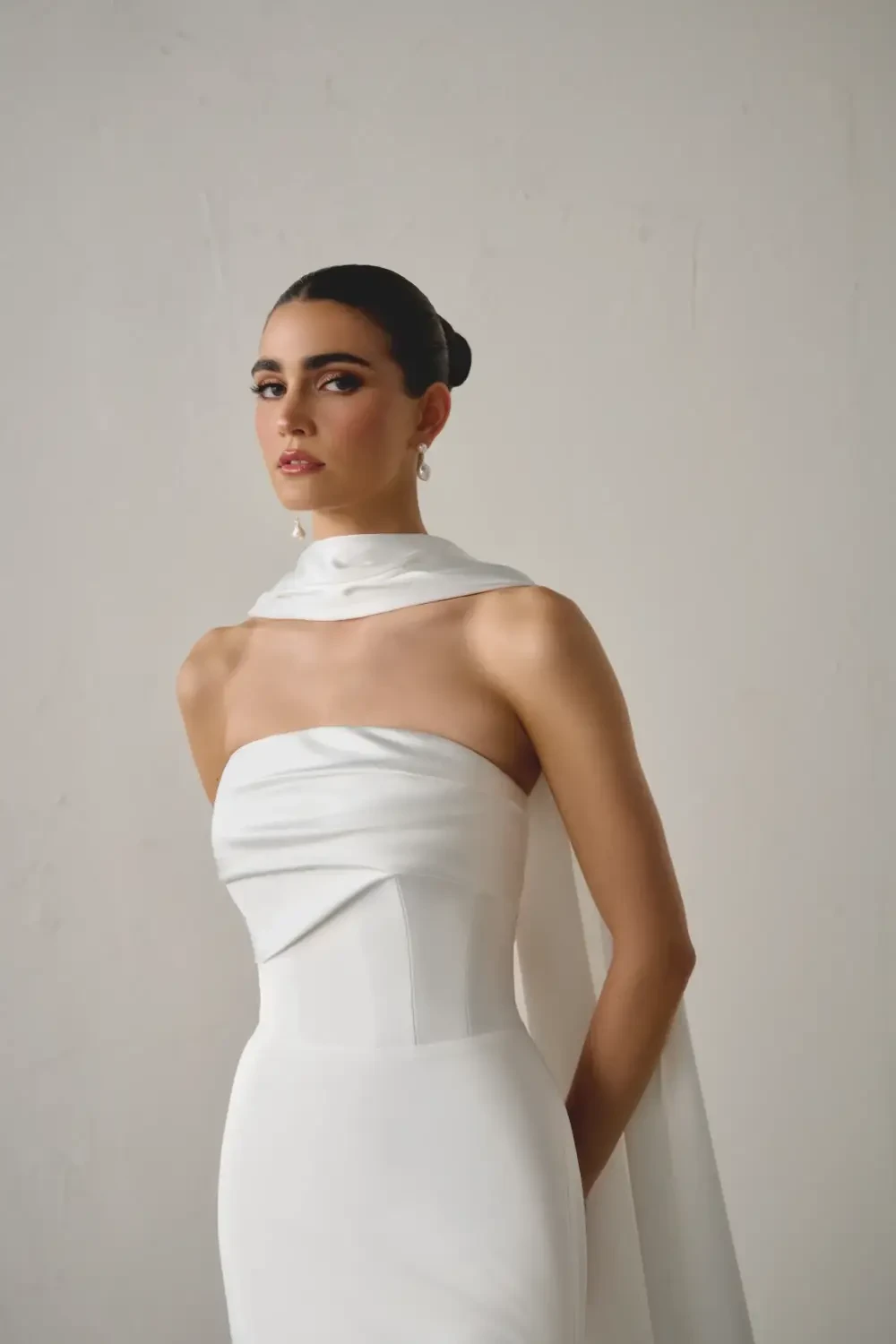A model wearing a sleek white strapless gown with a draped scarf around the neck, posing against a neutral background.