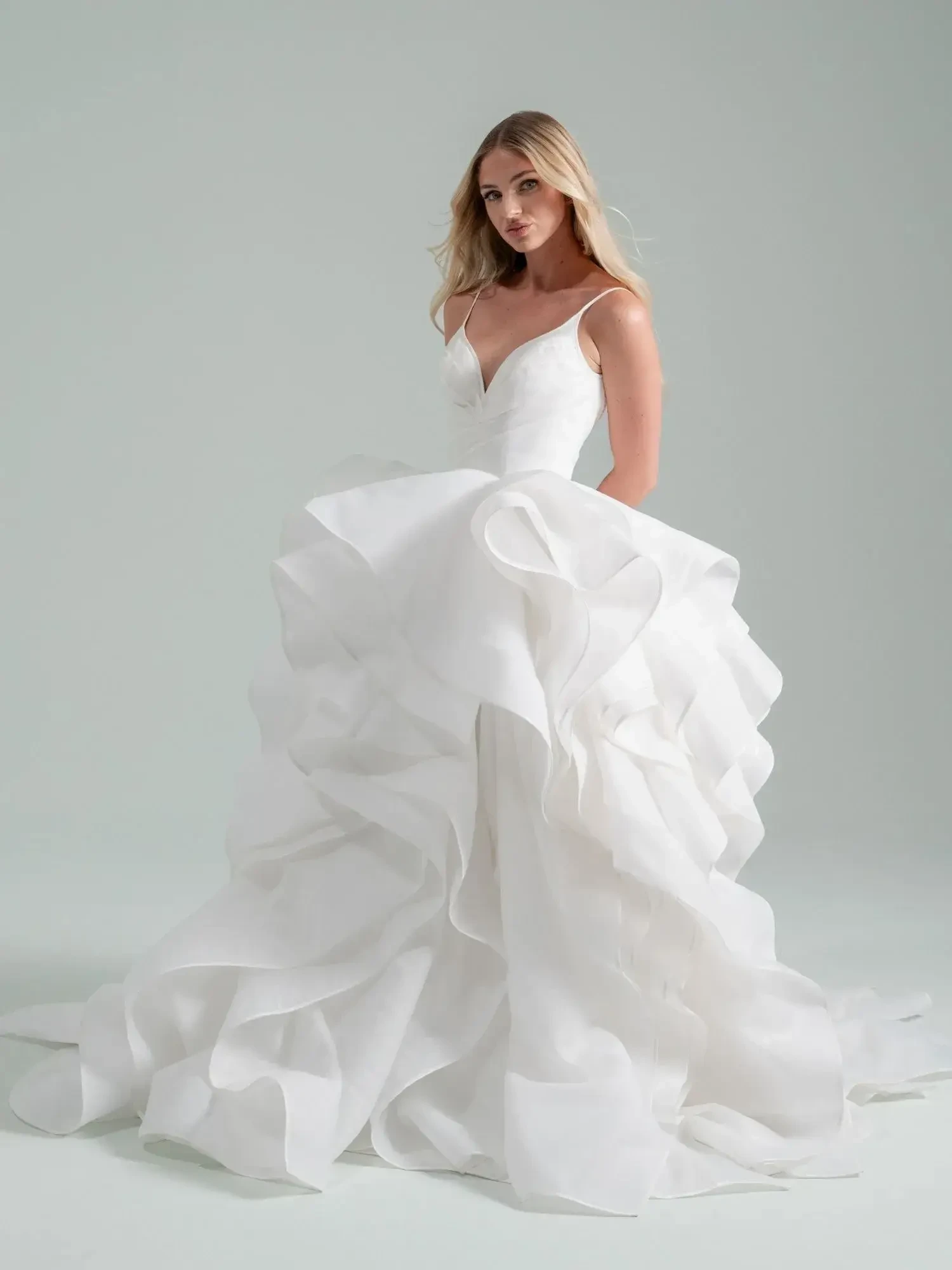 A woman in a voluminous white wedding gown featuring layered ruffles and a fitted bodice poses against a neutral background, exuding elegance.