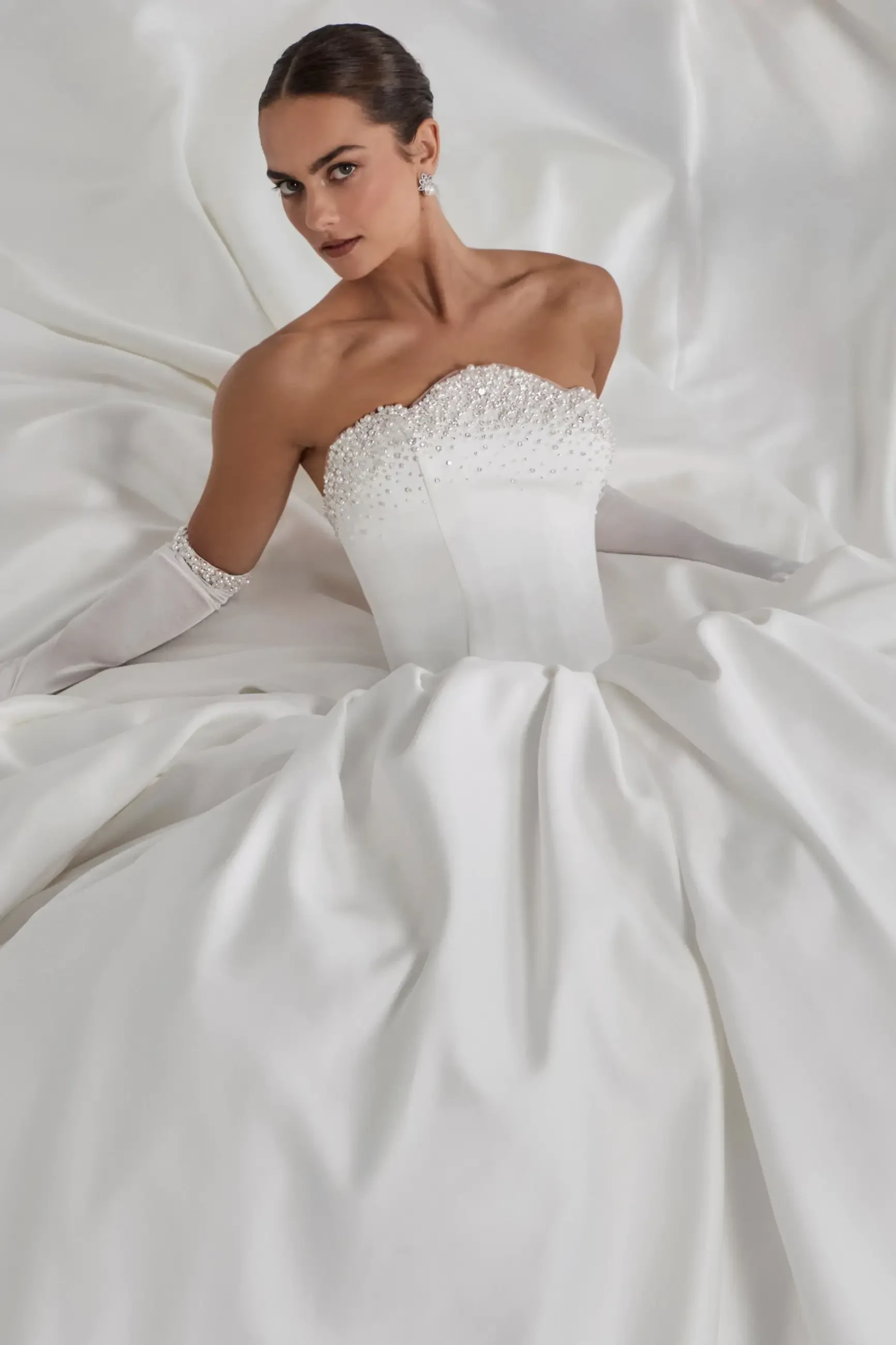 A woman in a strapless white gown with intricate beading, posing gracefully against a background of flowing white fabric.