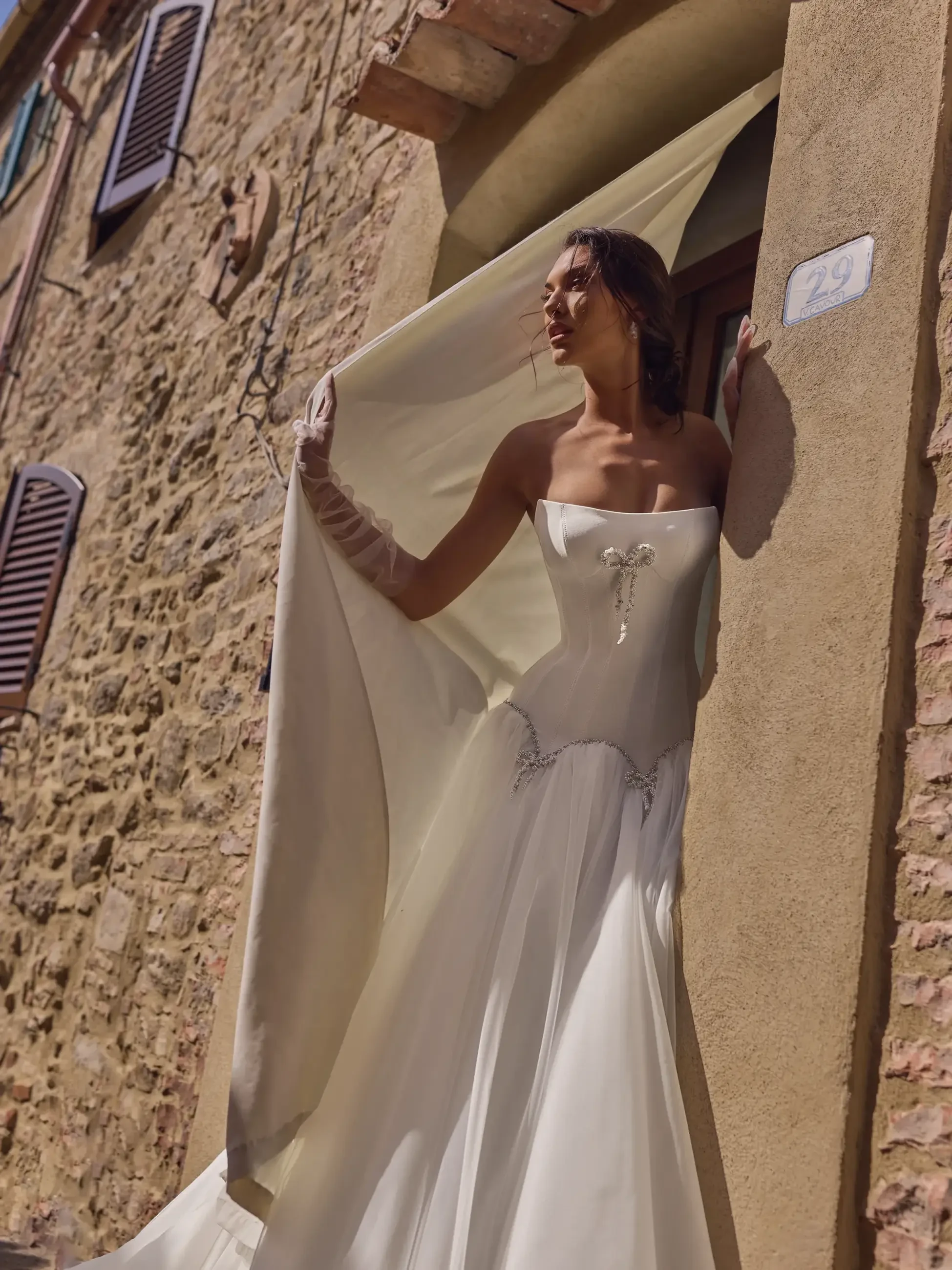 A bride in a stylish white wedding dress with a flowing cape stands gracefully by the entrance of a rustic stone building.