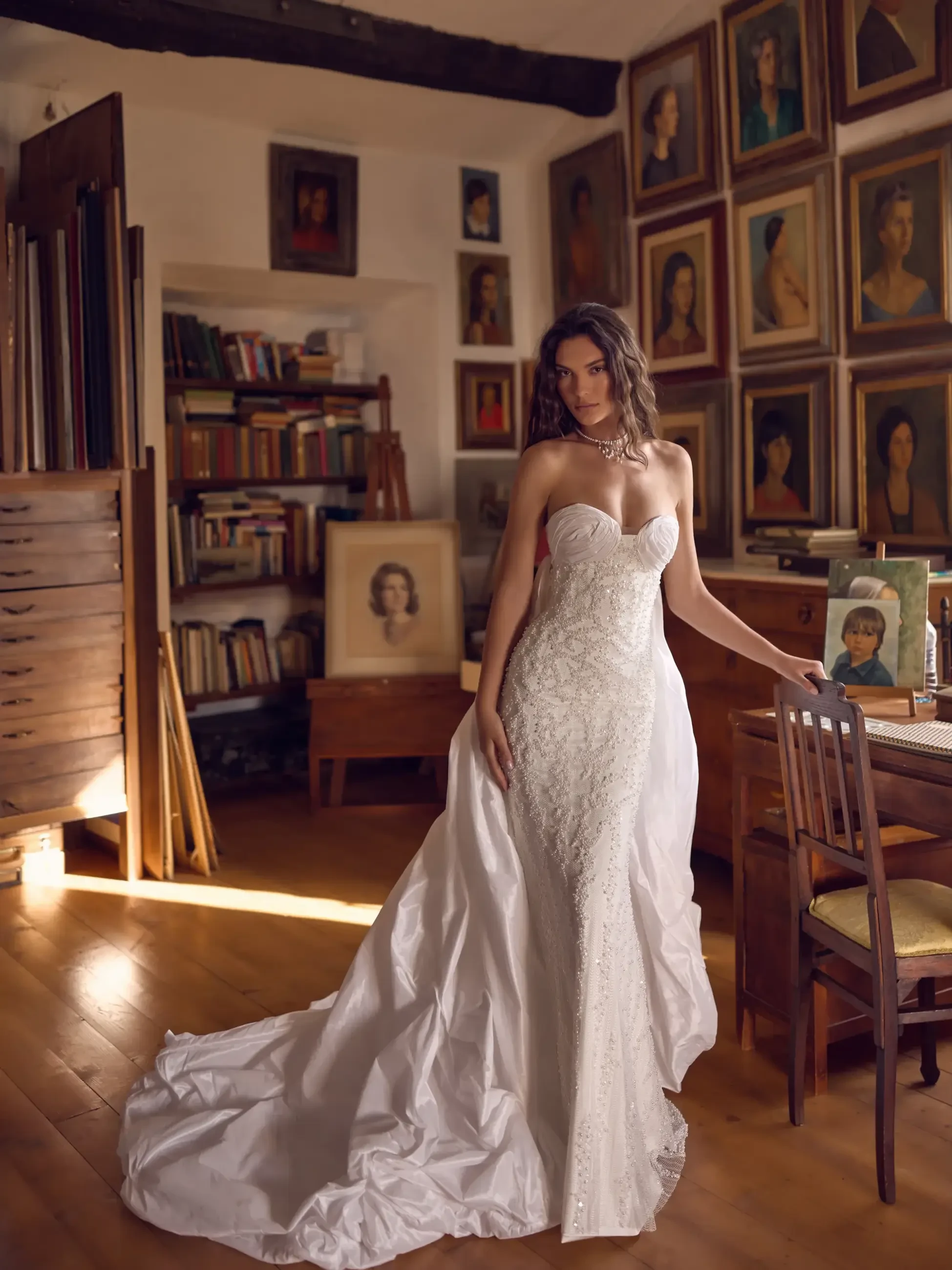 A woman in a white wedding dress stands in a room decorated with several framed portraits on the walls, surrounded by wooden furniture and bookshelves.
