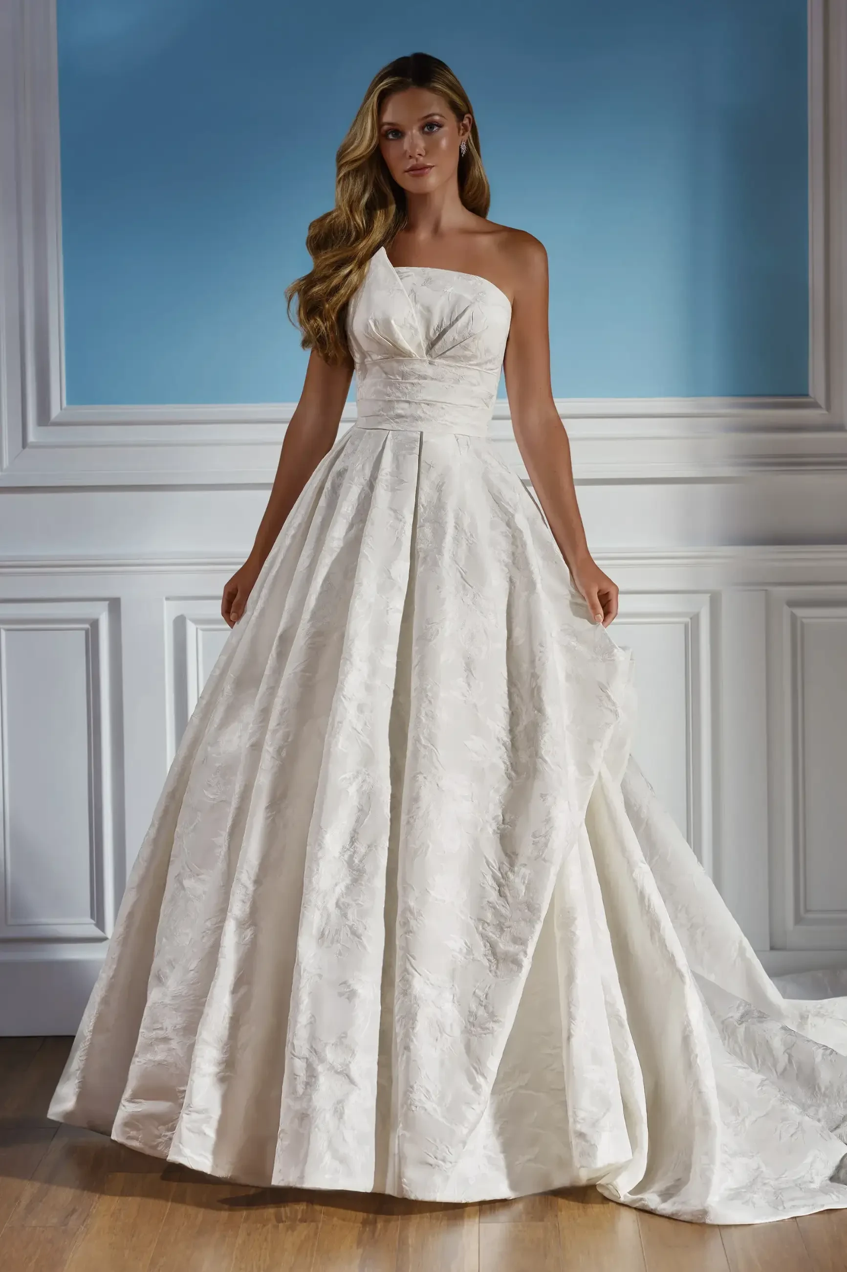 A woman stands in an elegant, strapless white bridal gown with a full skirt and subtle floral pattern. The setting is a softly lit room with blue walls.