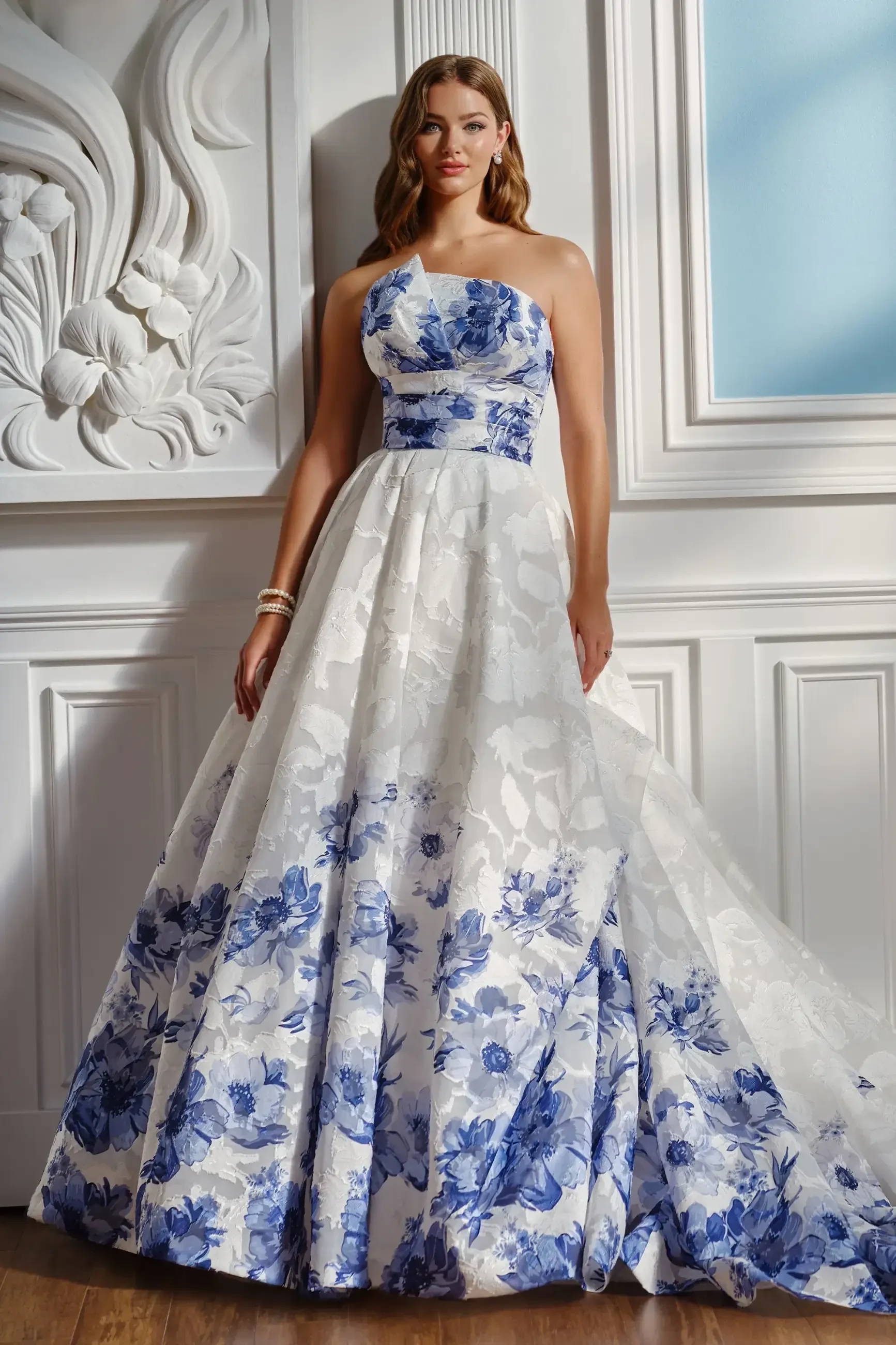 Woman in a strapless ball gown with blue floral patterns on a white background, standing against an ornate white interior wall, exuding elegance.