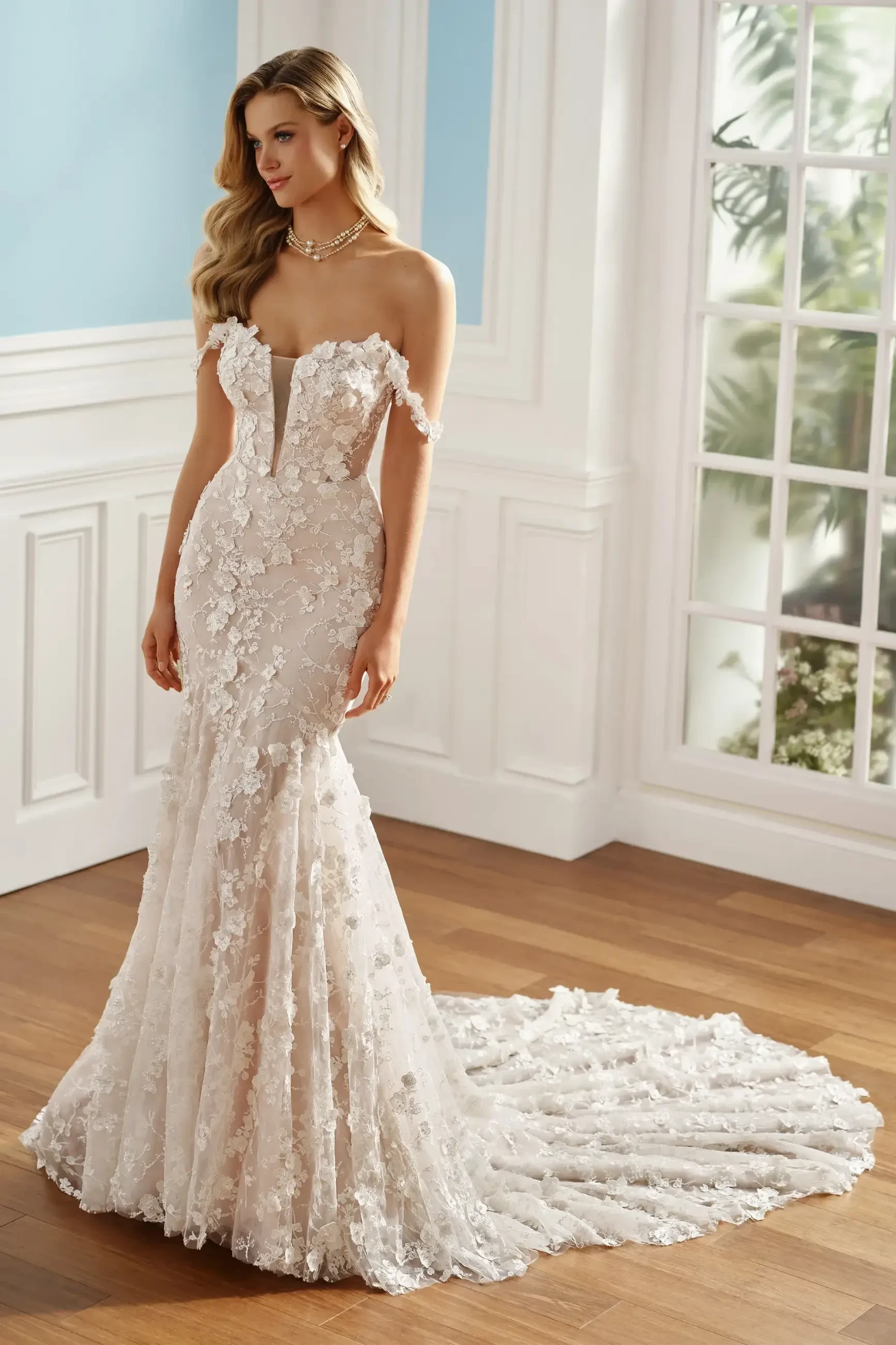 A woman in a white, floral lace wedding gown stands in a bright room with wooden floors and large windows, exuding elegance and serenity.