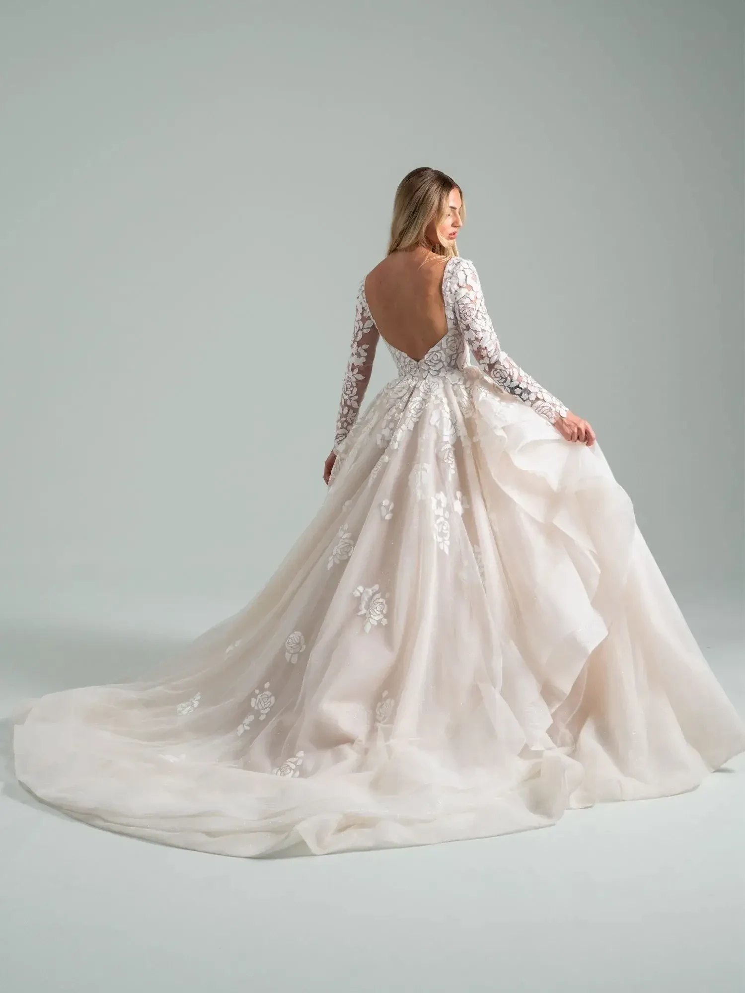 A woman in a flowing wedding dress with long sleeves and an open back, standing gracefully with her back to the camera. The gown features intricate lace detailing and a train.